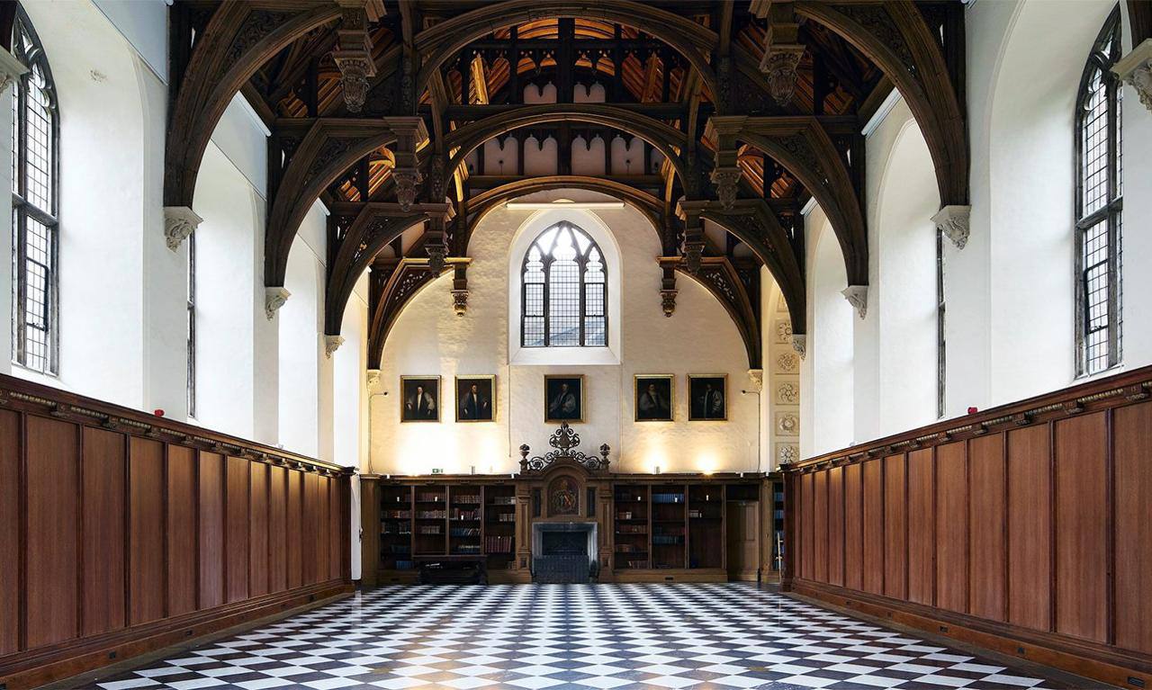 Spacious Gothic-style hall with carved wooden roof arches, panelled walls, a fireplace, bookcases and portraits. The floor is black and white tile.