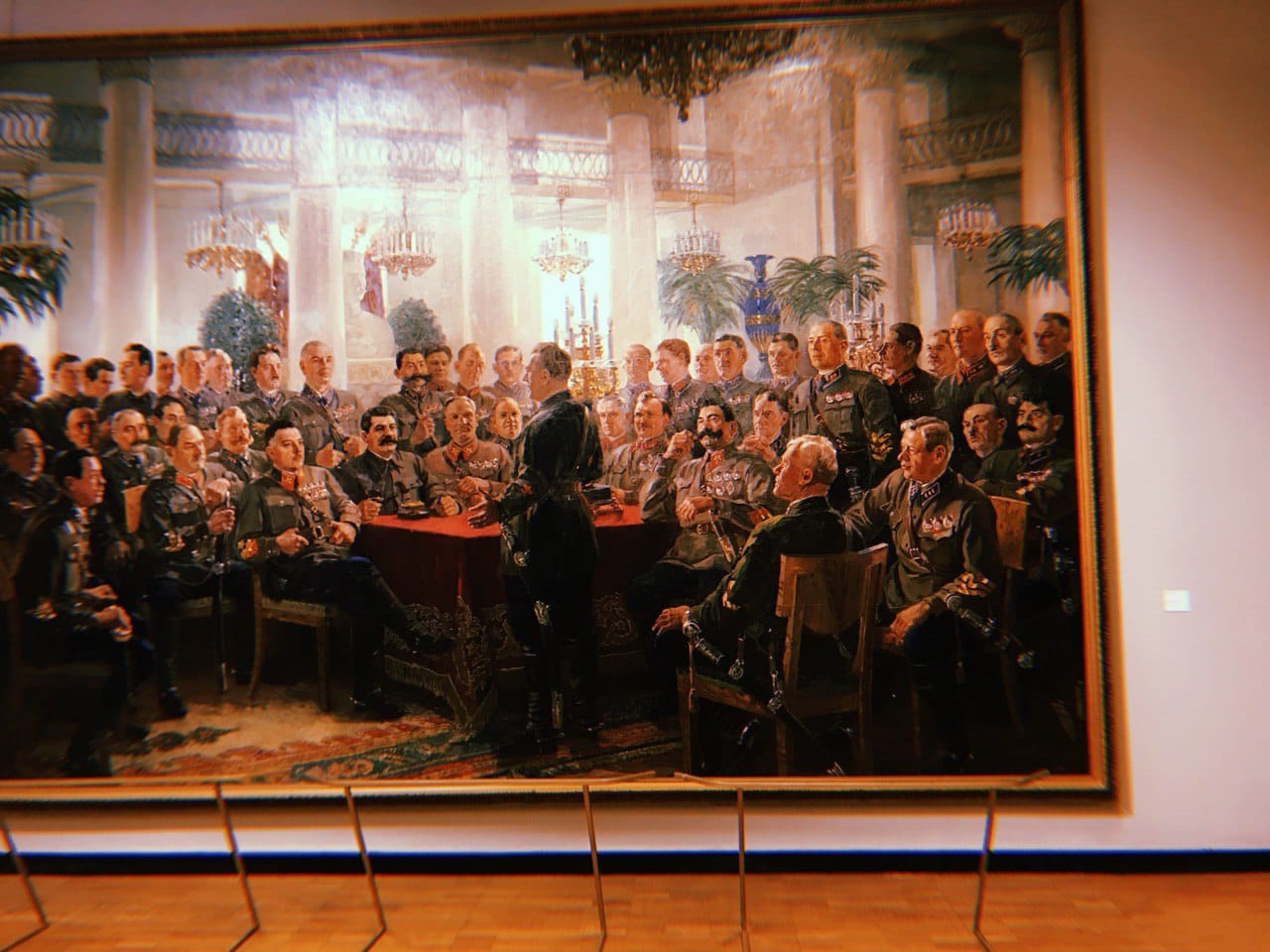 A large painting in a museum hall: uniformed military men around a table with a red cloth in a room with columns and chandeliers.
