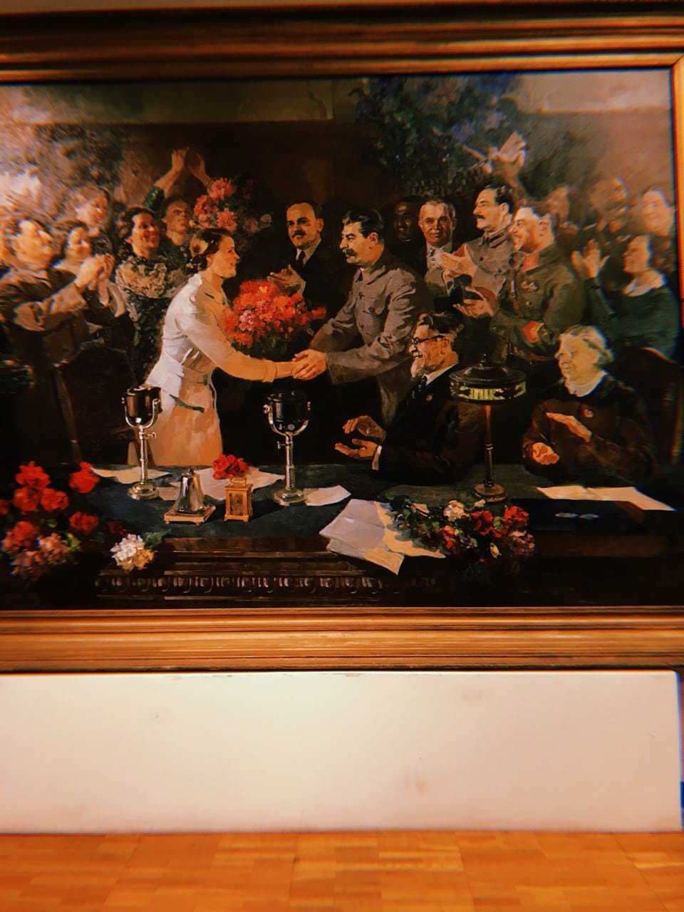 Grand tableau encadré : une jeune femme en robe claire, avec un bouquet rouge, serre la main d'un homme en tunique grise, entourée de gens qui applaudissent ; micros et fleurs sur la table.