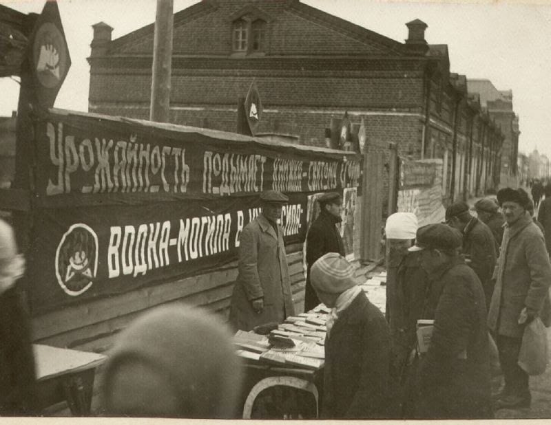 Men in coats and caps browse books on a street stall. A long banner about harvests and the harm of vodka hangs on a fence; a brick building stands behind.