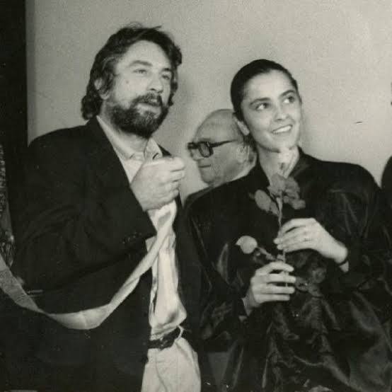 Photo en noir et blanc : un homme barbu aux cheveux sombres, en veste, près d'une jeune femme souriante tenant une rose. Un homme âgé à lunettes se tient derrière eux.