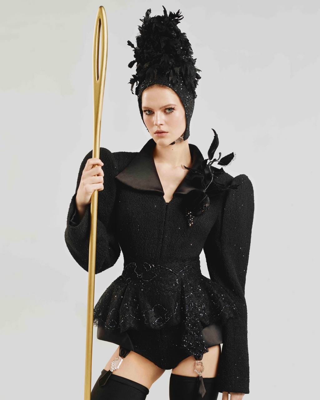 A young woman in a black bouclé jacket with a lace peplum, stockings and a feathered cap holds an oversized golden needle against a light grey background.