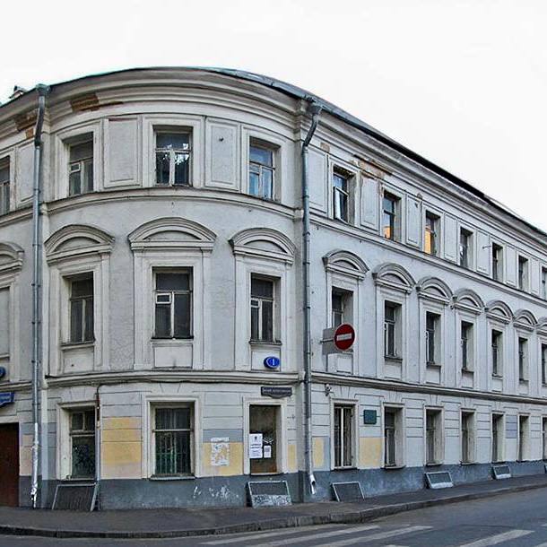 Ancien immeuble d'angle enduit à façade arrondie, avec fenêtres à encadrements cintrés et gouttières. Un trottoir et un panneau de sens interdit sont à côté.