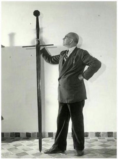 Sur une photographie en noir et blanc, un homme en lunettes et costume se tient près d'une énorme épée plus haute que lui.