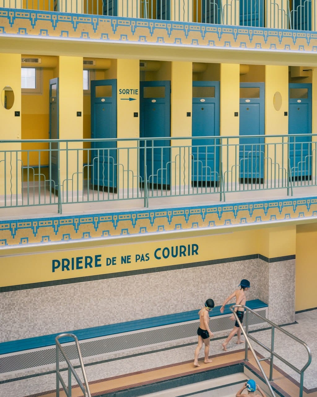 Deux garçons en bonnet de bain marchent au bord du bassin. Au-dessus, des galeries jaunes à cabines bleues, une frise en mosaïque et l'inscription « Prière de ne pas courir ».