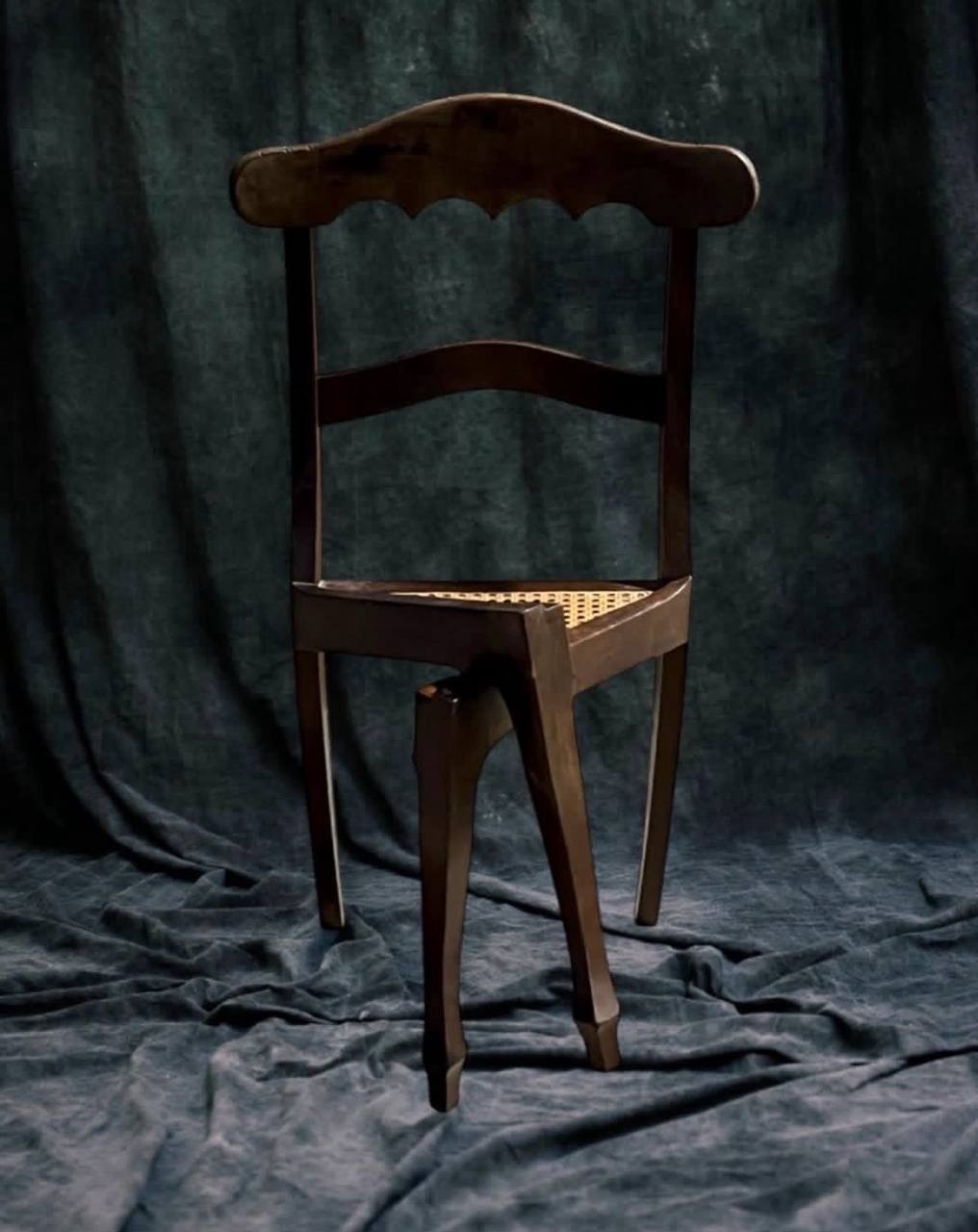 A dark wooden chair with a woven cane seat and curved front legs against a dark draped backdrop.