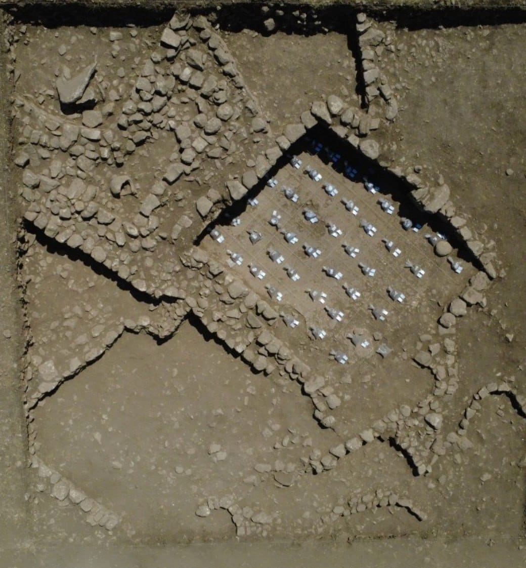 Vue du dessus d'une tranchée archéologique aux murs de pierre irréguliers. Au centre, une zone rectangulaire quadrillée avec des rangées de petits repères clairs.