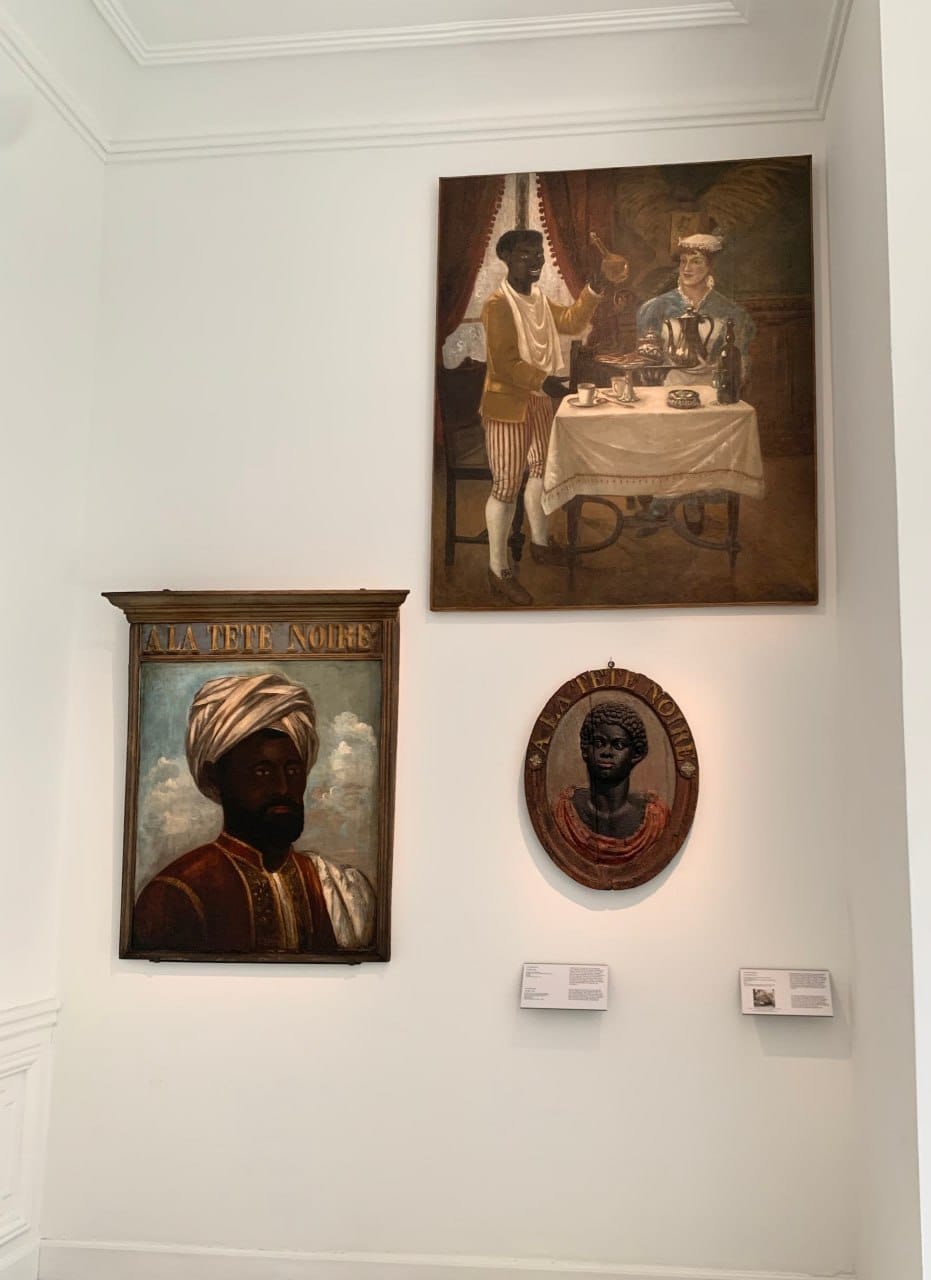 Trois œuvres sur un mur blanc : deux enseignes « A la Tête Noire », un portrait d'homme en turban et un buste en relief ovale, et un tableau d'un serviteur près d'une table dressée.