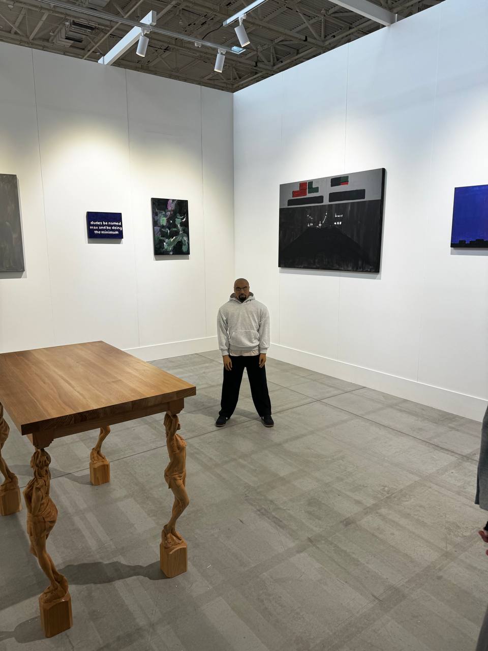 In a white gallery room hang abstract paintings, one with a text line. A wooden table on figure-shaped legs stands in front; a figure of a man in a gray hoodie and black trousers is in the c