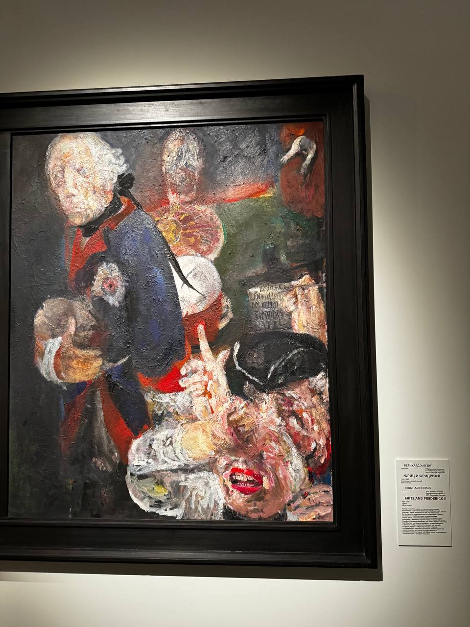 Expressive thick-brushed painting in a dark frame: a white-haired man in a blue and red uniform, beside a distorted face with red lips and a raised finger. A wall label is at right.