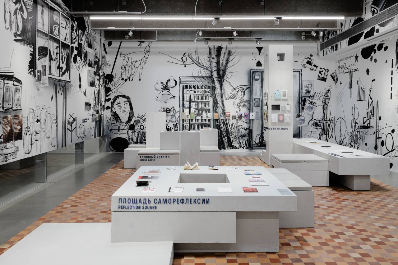 Hall with white walls covered in black drawings of a tree, birds and faces. Concrete tables with books stand on a mosaic tile floor; one reads «Reflection Square».