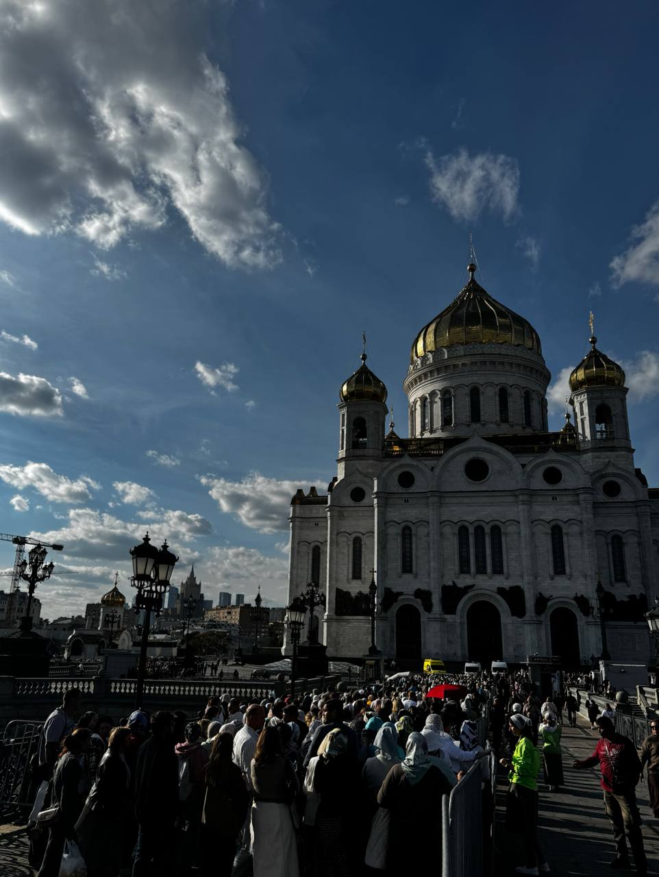A white-stone cathedral with golden domes against a deep blue sky with clouds. In the foreground, a dense crowd, some women in headscarves, stands by metal barriers and street lamps.