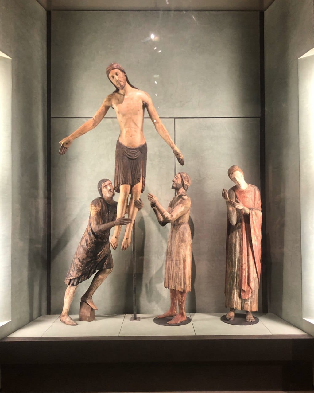 Dans une vitrine de musée, quatre figures en bois polychrome : le Christ aux bras écartés et trois personnages à ses côtés.