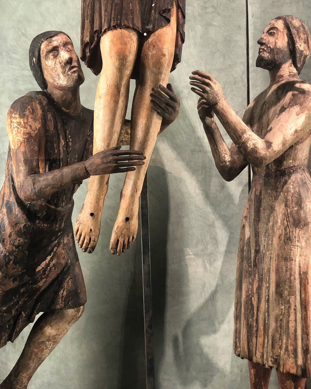 Deux anciennes figures en bois peint soutiennent les jambes d'un crucifié, aux pieds percés de trous de clous. La peinture est usée, le fond gris-vert pâle.