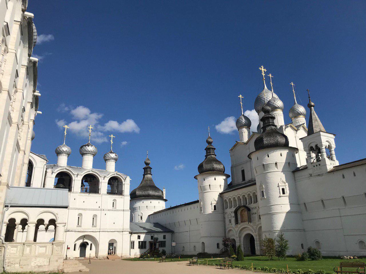 Murs et tours blancs aux toits sombres en pointe, église aux bulbes argentés et croix dorées, clocher à gauche. Ciel bleu au-dessus de la cour, pelouse et bancs au premier plan.