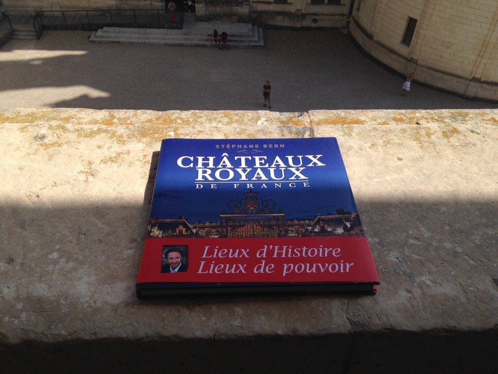 Le livre « Châteaux royaux de France » de Stéphane Bern posé sur un parapet de pierre au-dessus d'une cour où l'on voit quelques personnes.