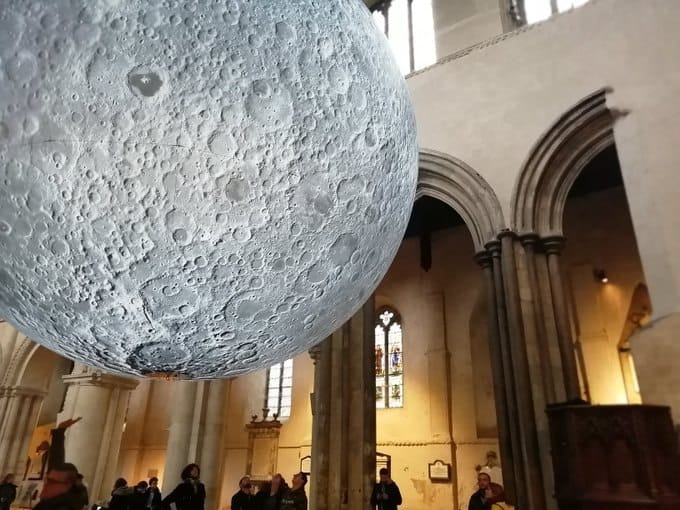 Une énorme sphère à la surface lunaire détaillée est suspendue dans une église aux arcs de pierre et aux vitraux. Des visiteurs l'observent d'en bas.