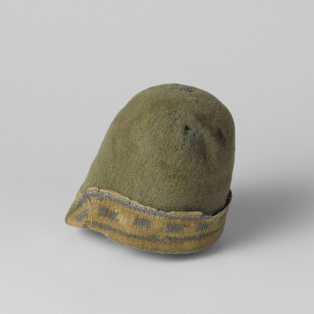 Bonnet conique en feutre vert, usé et percé de petits trous. Le bord inférieur est relevé et montre une bande jaune ornée d'un motif géométrique gris-bleu. Fond gris clair.