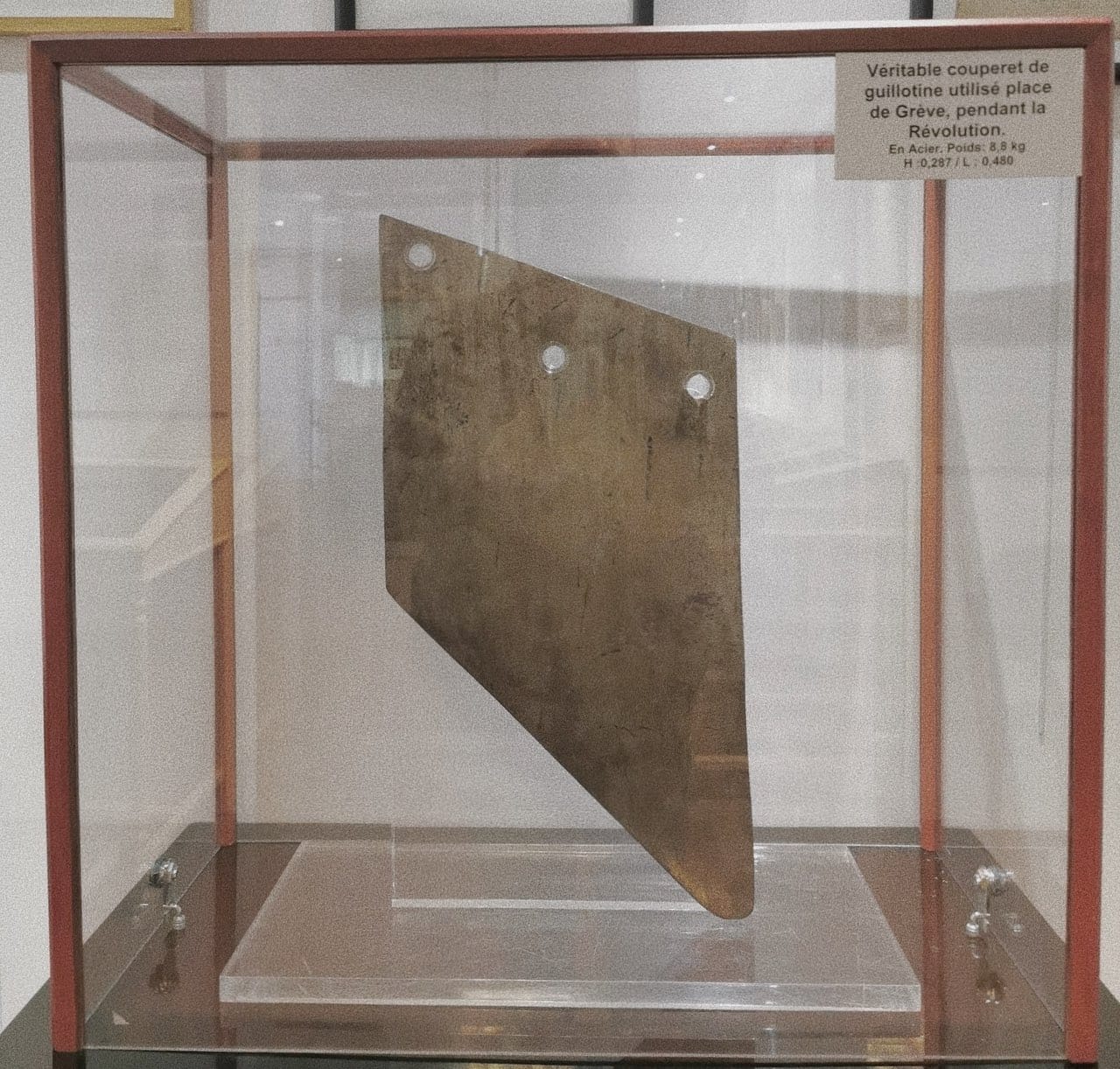 A wide steel guillotine blade with three holes in a glass display case with a French label.