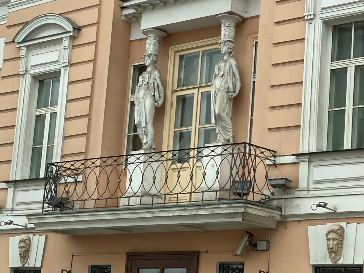 Deux cariatides blanches encadrent la porte d'un balcon à garde-corps en fer forgé. La façade est pêche, avec des fenêtres à encadrements blancs et des mascarons de lion.