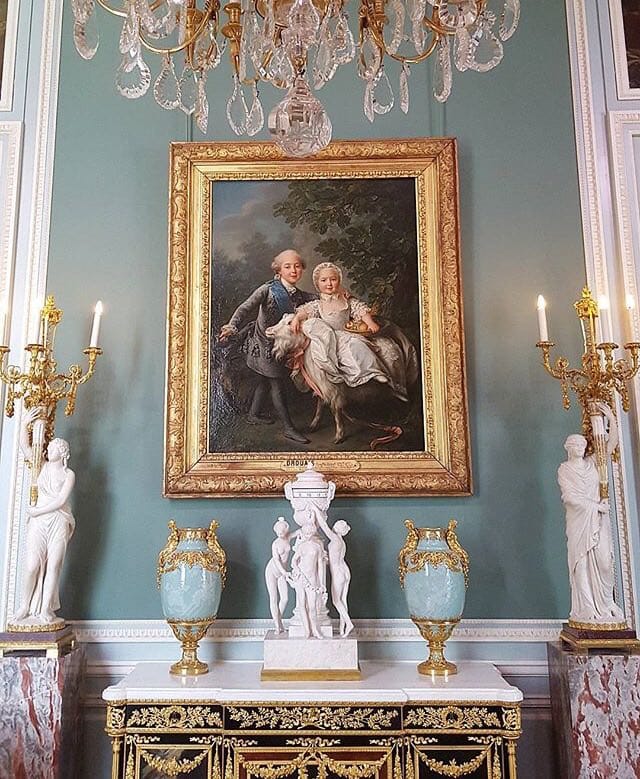 A children's portrait with a goat in a gilded frame on a turquoise wall under a chandelier, with a chest of drawers, vases and statuettes below.
