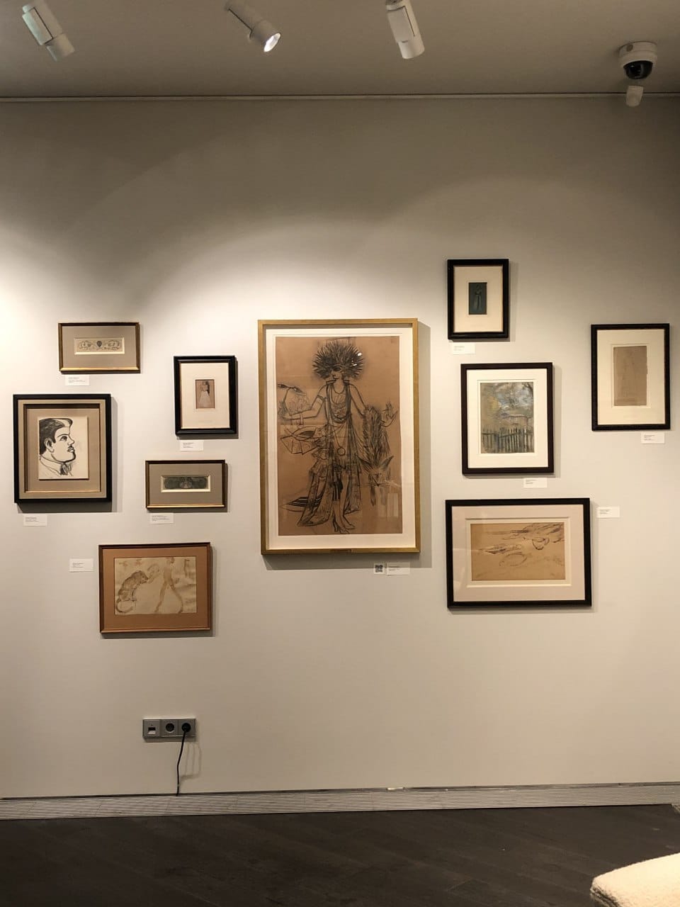 Gallery wall with a large sketch of a costumed female figure in a gold frame at the centre, surrounded by smaller drawings in dark frames.
