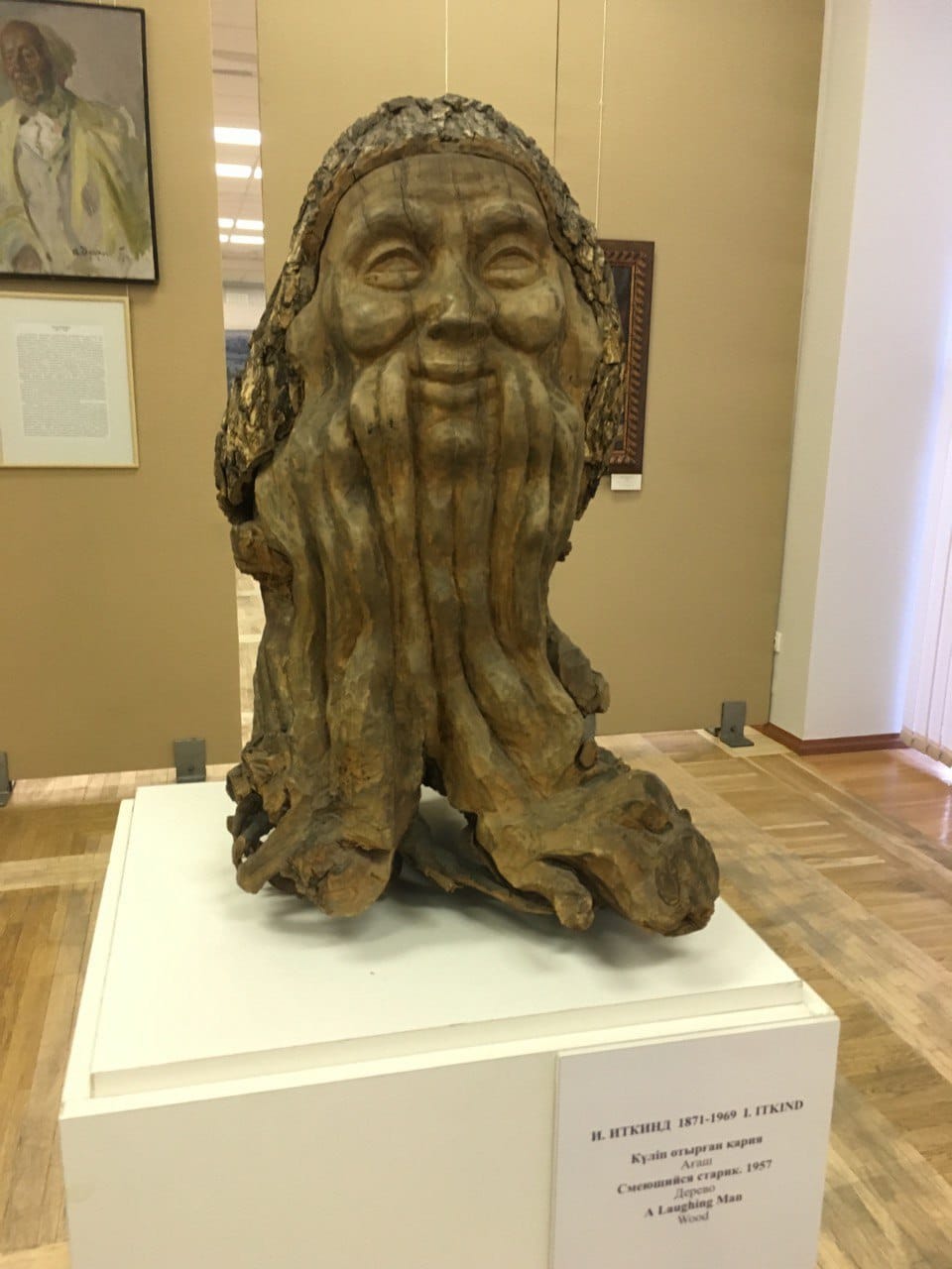 Sculpture en bois d'un vieillard souriant dont la longue barbe se fond dans des racines noueuses, sur un socle blanc avec cartel, dans une salle de musée.