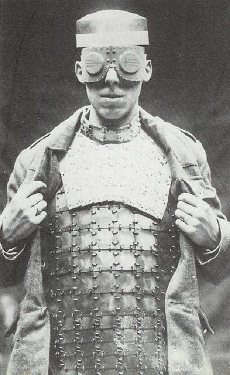 Black and white photo of a man in a cap with protective goggles and a plate breastplate worn under an open greatcoat.