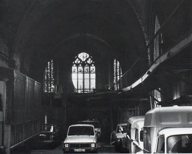 Photo en noir et blanc : voitures et camionnettes dans la nef sombre d'une église gothique, avec vitraux et échafaudages.