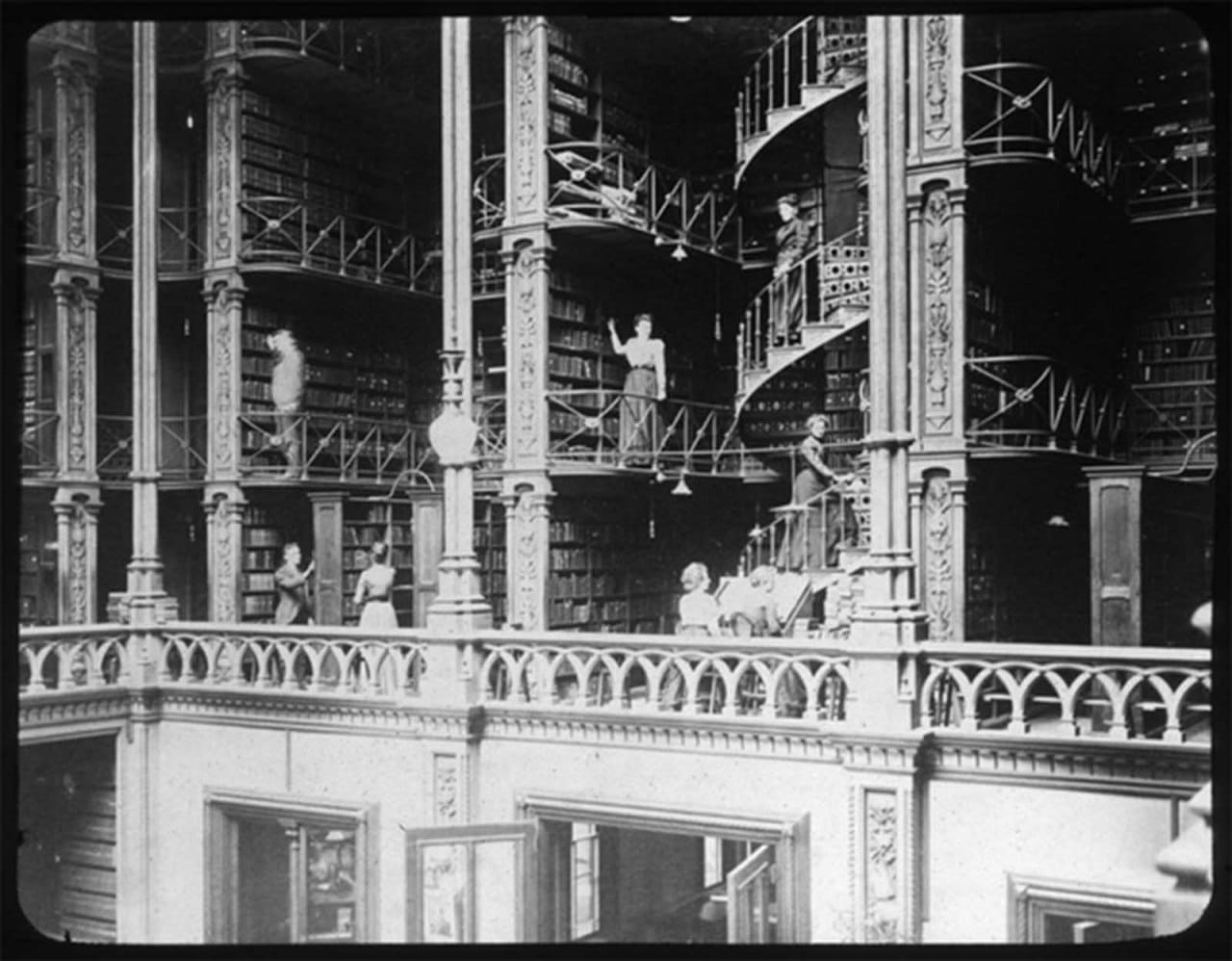 Ancienne photographie d'une bibliothèque : galeries en fonte ornées, livres sur plusieurs niveaux, escalier en colimaçon, femmes sur les balcons et lecteurs assis derrière une balustrade.