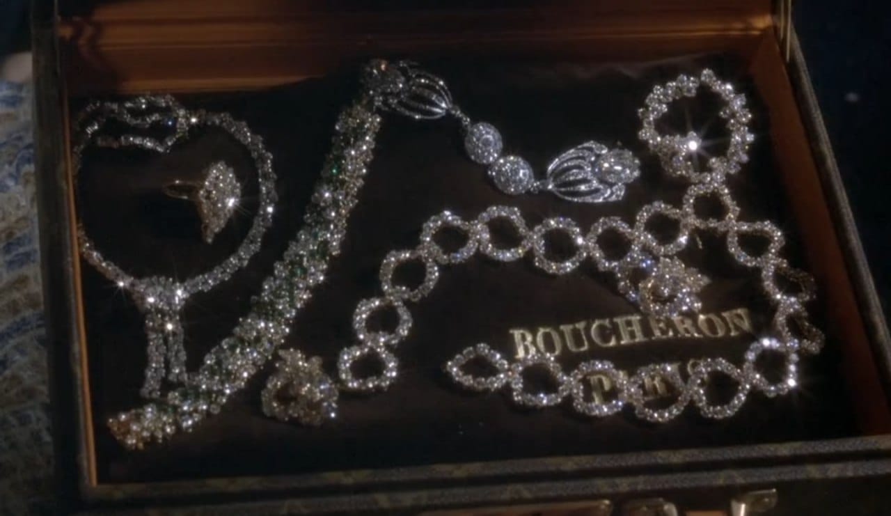 Un écrin ouvert, garni de velours sombre, contient colliers, bracelets, boucles d'oreilles et bagues en diamants, certains avec émeraudes. Le nom Boucheron figure sur le tissu.