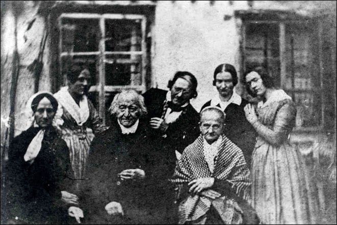 Old black-and-white group photograph of seven people by a house window, two elderly sitters in the centre.