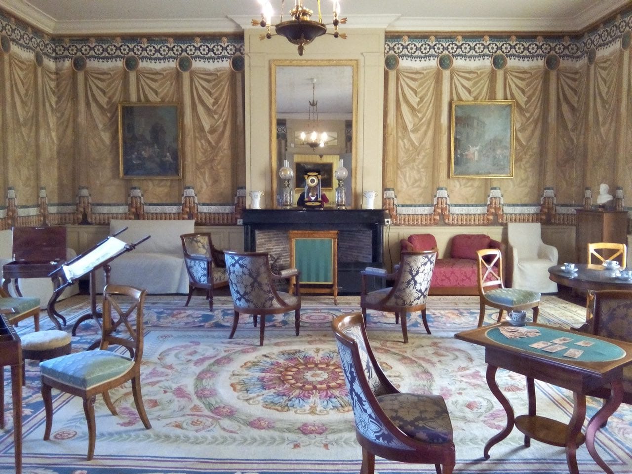 Salon du château de Saché aux murs peints en draperies, avec miroir au-dessus de la cheminée, fauteuils, table de jeu et tapis à motifs floraux.