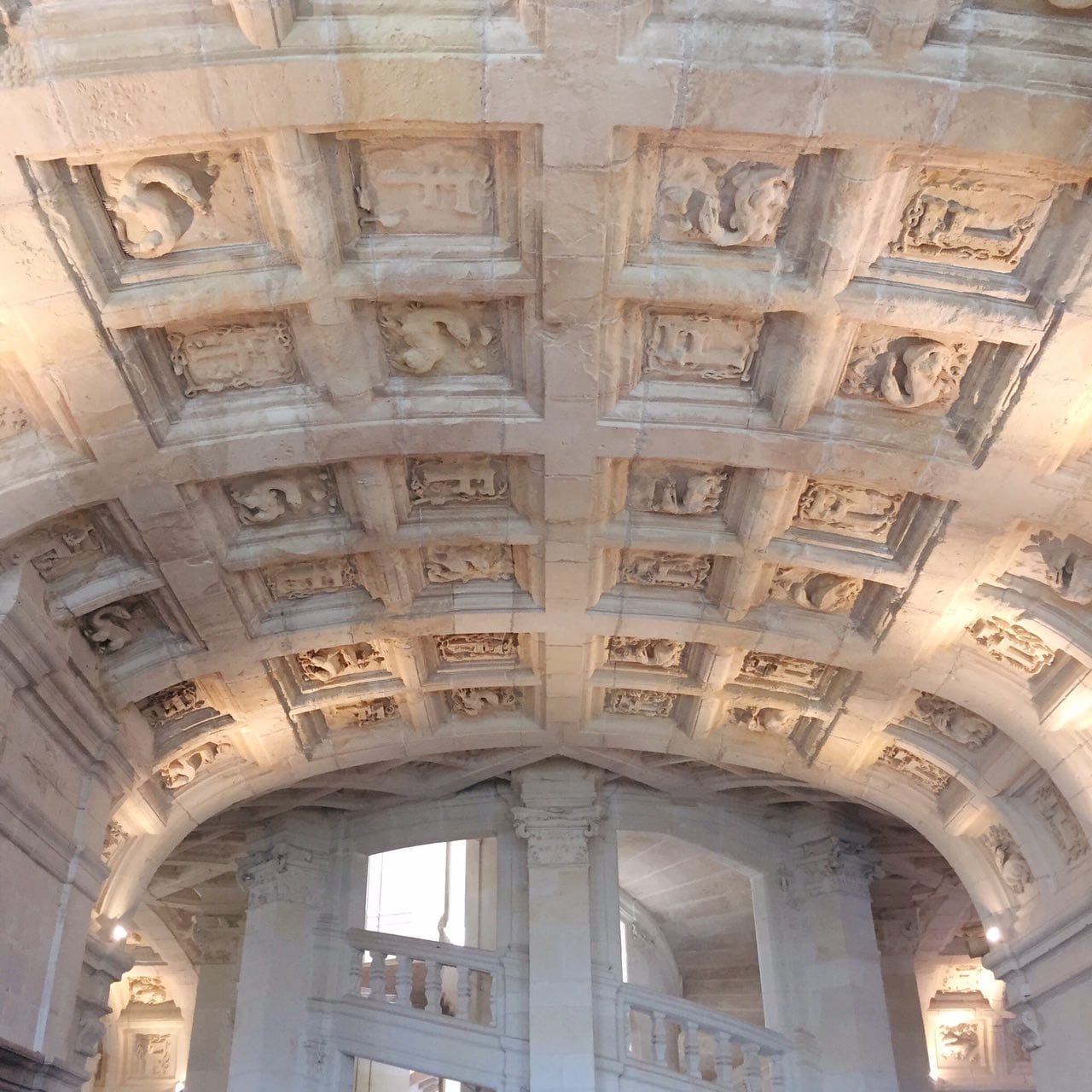 Stone coffered vault of a gallery with reliefs of salamanders and the letter F, with a balustraded staircase visible below.