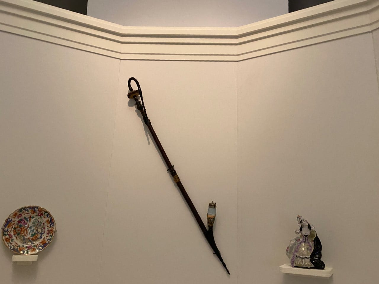 Une longue pipe à tuyau courbe et fourneau peint est accrochée en diagonale sur un mur clair. À gauche, une assiette en porcelaine fleurie ; à droite, une statuette de dame.