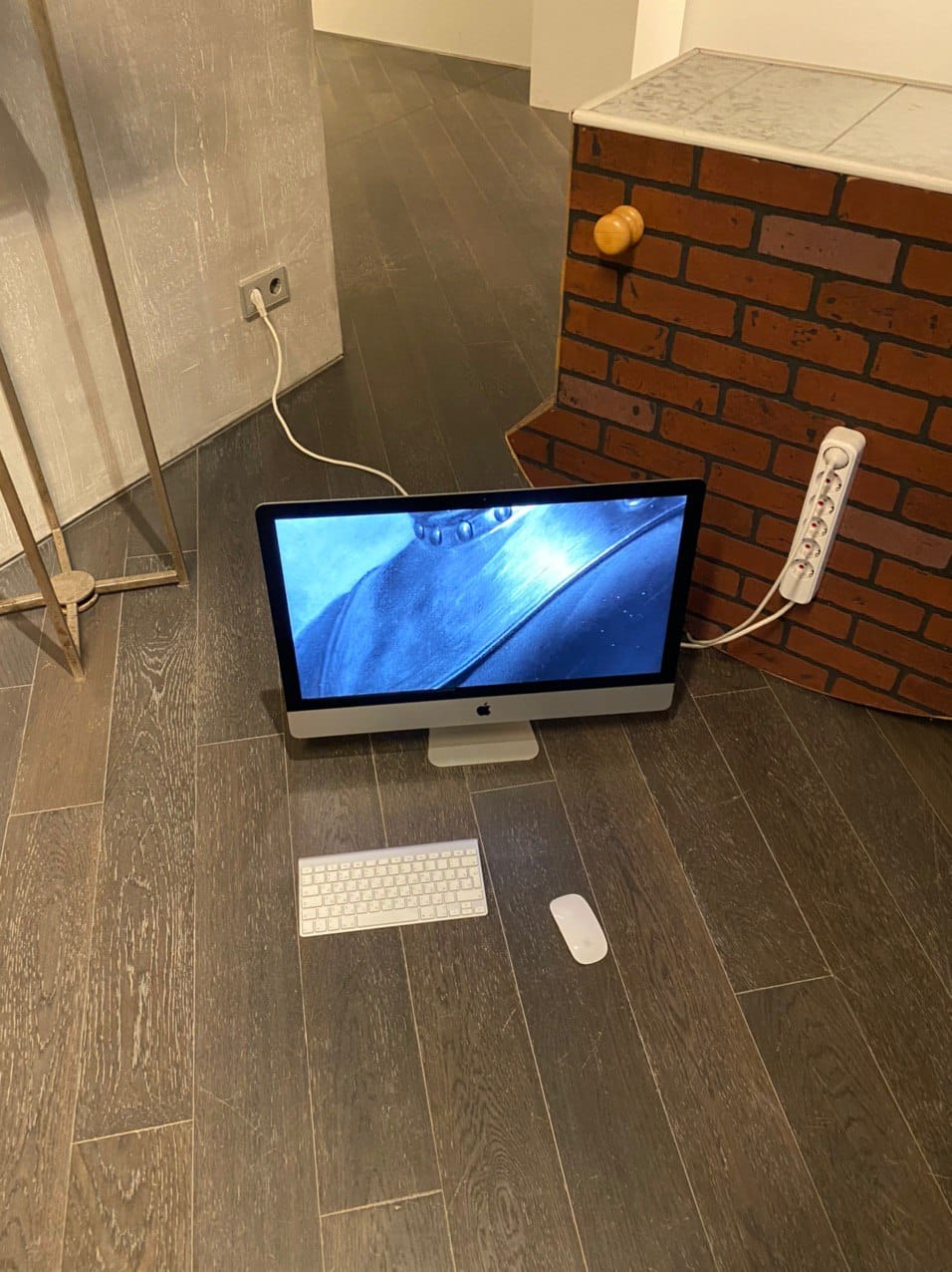 Sur un parquet sombre, un ordinateur tout-en-un affiche une image vidéo bleue, avec clavier et souris. À côté, un objet en faux briques et une multiprise.