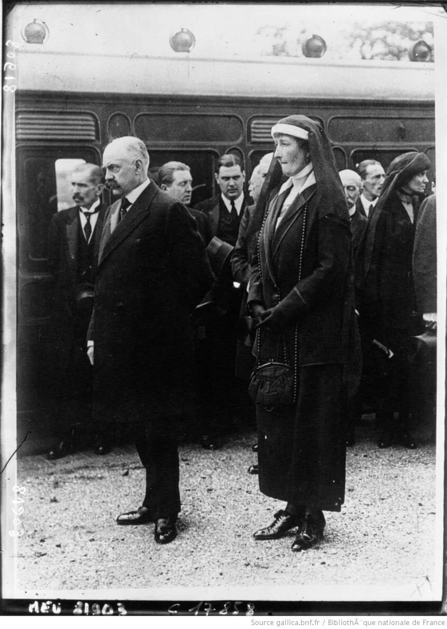 Photo en noir et blanc : un homme âgé en manteau et une femme voilée de sombre devant un train, avec un groupe d'hommes en costume sombre derrière.