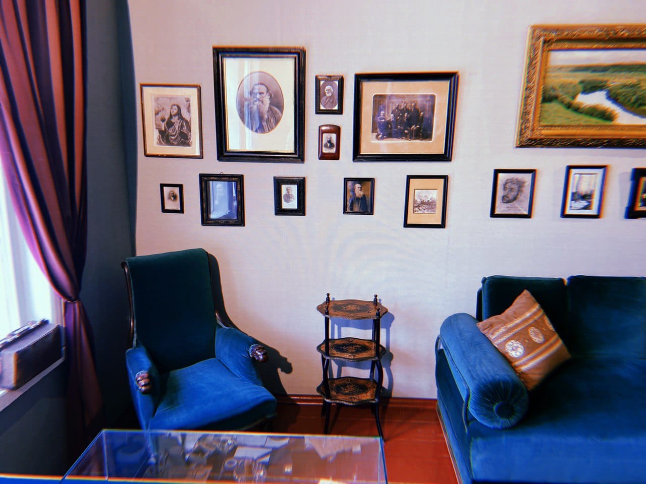 Un mur orné de portraits en noir et blanc encadrés et d'un paysage dans un cadre doré. En bas : fauteuil et canapé en velours, guéridon à trois plateaux, vitrine et rideau.