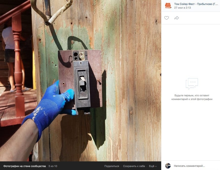 Une main gantée de bleu tient un vieux disjoncteur fixé sur un panneau sombre, sur une planche aux restes de peinture verte. À gauche, la rampe d'un perron.