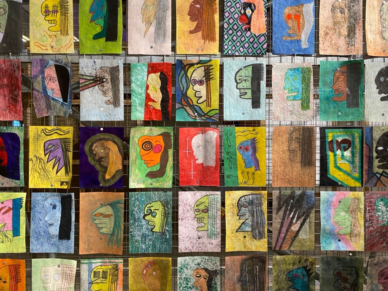 Rows of colourful drawn heads in profile on sheets of paper fixed to a metal grid.