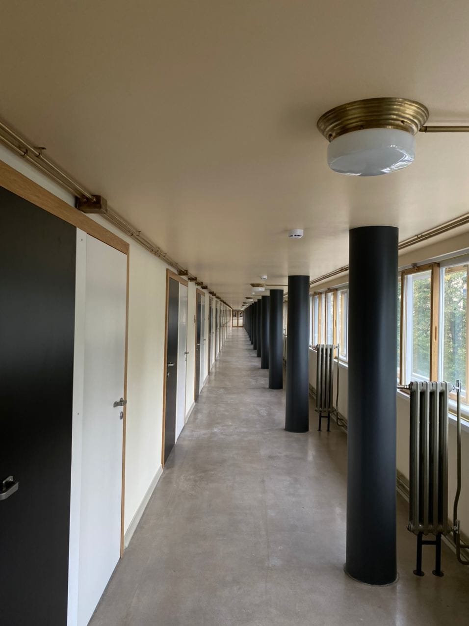 A long corridor with black and white doors on the left, dark columns, brass ceiling lights, and windows with radiators on the right.