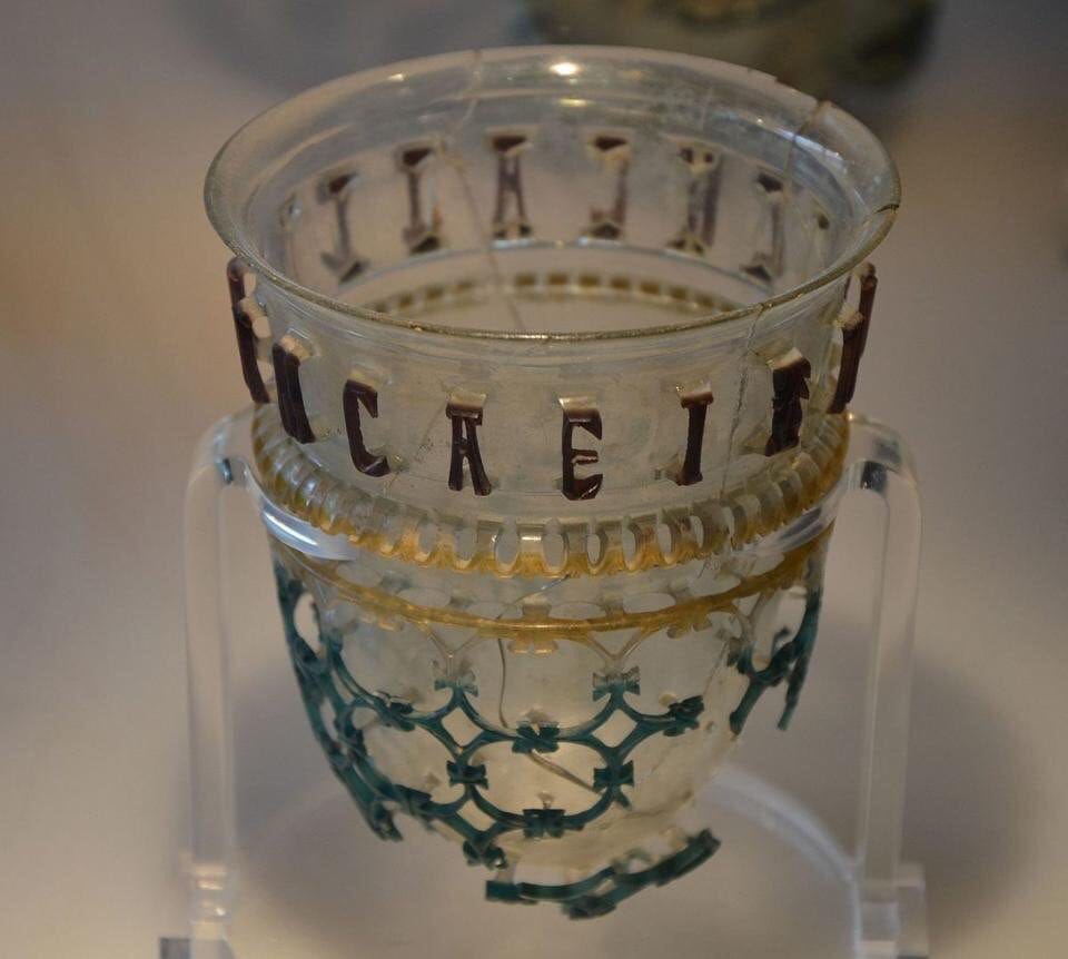Gobelet romain en verre transparent, avec une résille verte ajourée en bas et les lettres sombres d'une inscription autour du bord.