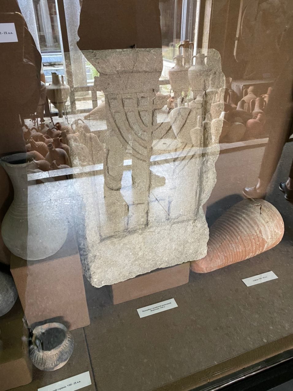 Dalle de calcaire ornée d'une menora à sept branches en relief, dans une vitrine de musée. Une amphore et des cruches en céramique sont à côté ; des reflets sur la vitre.
