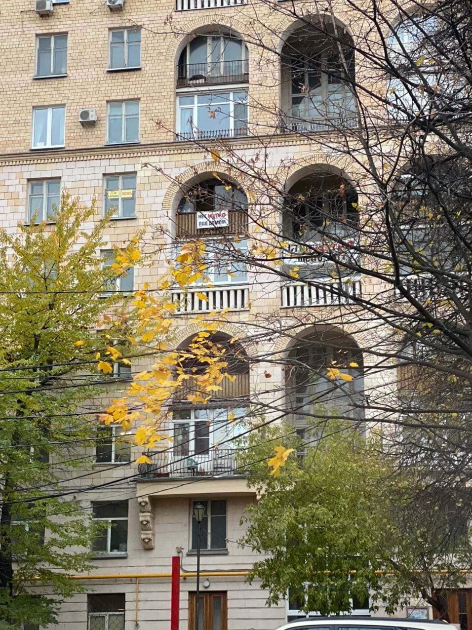 Façade en brique d'un immeuble à loggias en arc derrière des arbres d'automne ; une banderole sur un balcon dit « Нет метро под домом » (pas de métro sous la maison).