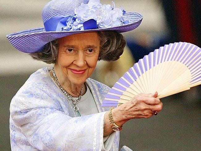 Une femme âgée souriante, coiffée d'un chapeau lilas à fleurs et vêtue d'une veste claire à motifs, tient un éventail ouvert. Elle porte un collier et un bracelet doré.
