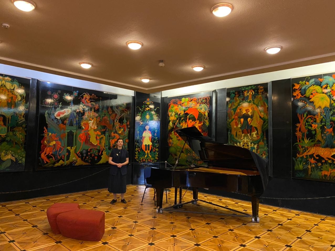 Un piano à queue noir se trouve dans une salle au parquet, près d'une femme en tenue sombre. Les murs sont ornés de panneaux laqués colorés à motifs de contes ; deux poufs rouges au sol.