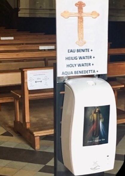 Distributeur automatique blanc avec une croix et la mention « eau bénite » en quatre langues, orné d'une image du Christ, dans une église près de bancs en bois.
