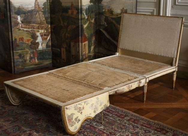 Lit de repos en bois à haut dossier, doré, avec tissu à fleurs sur la ceinture; l'assise est en toile brute. Derrière, un paravent paysager; au sol, tapis et parquet.