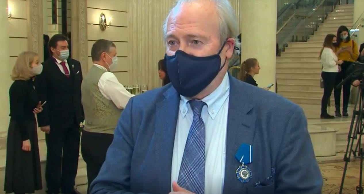 A man in a blue jacket and dark face mask, with a blue decoration on his lapel, stands in a hall with a wide staircase.