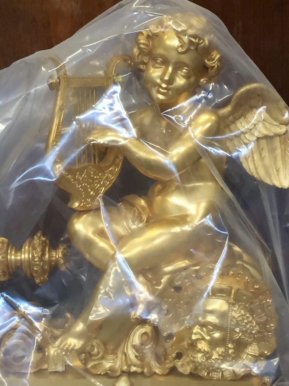 A gilded figure of a winged putto holding a lyre, wrapped in a transparent plastic bag.